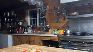 Une cuisine avec des armoires en bois, une cuisinière et des casseroles suspendues. On aperçoit une planche à découper avec des légumes sur le comptoir.