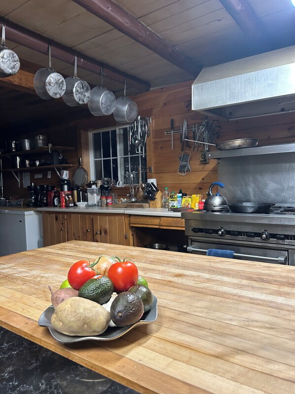 Una cocina con armarios de madera, una estufa y ollas colgantes. Hay una tabla de cortar con verduras sobre la encimera.