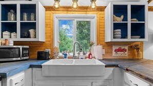 Eine Küche mit weißen Schränken, einer doppelten Spüle und Holzelementen. Regale mit Küchenutensilien und ein Fenster mit Blick auf Bäume.