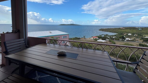 A balcony with a wooden table and bench, overlooking a coastal view with a clear sky and distant land.