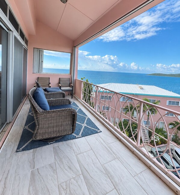 A balcony with wicker furniture, a view of the ocean, and a pink building in the background.