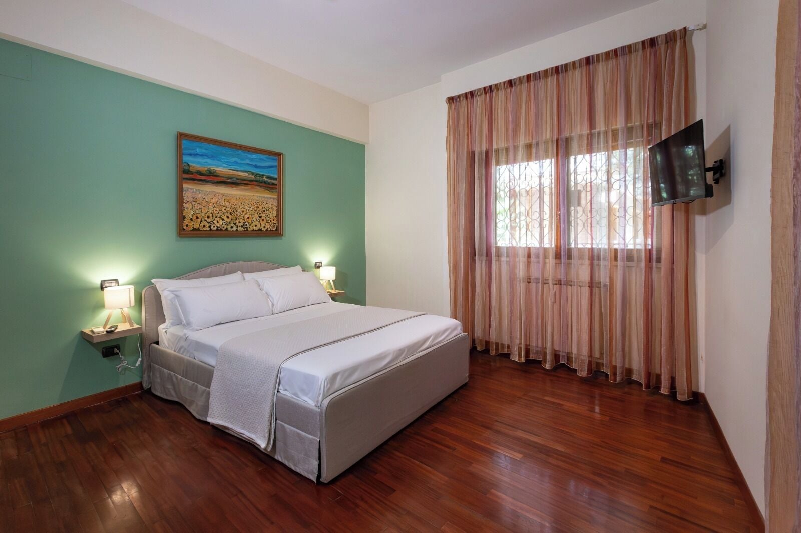 A bedroom with a bed, a painting on the wall, a TV mounted on the wall, and a window with curtains.