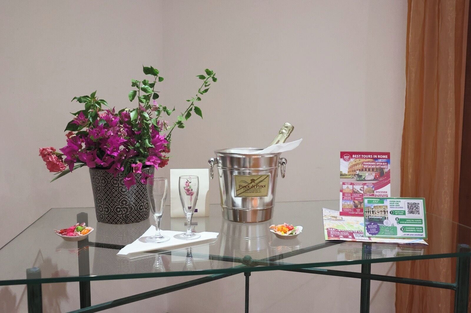 A glass table with a metal bucket labeled "Pinot Grigio," a vase with purple flowers, two glasses, a small plate with food, and a pamphlet titled "Best Tours in Rome."
