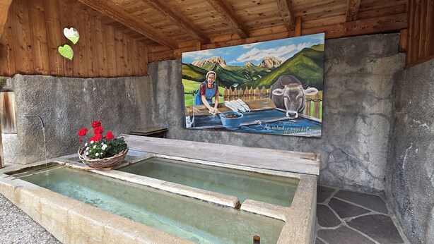 A rustic wooden structure with a stone water basin and a painting of a woman and cow. - Tamion House (Vigo di Fassa)