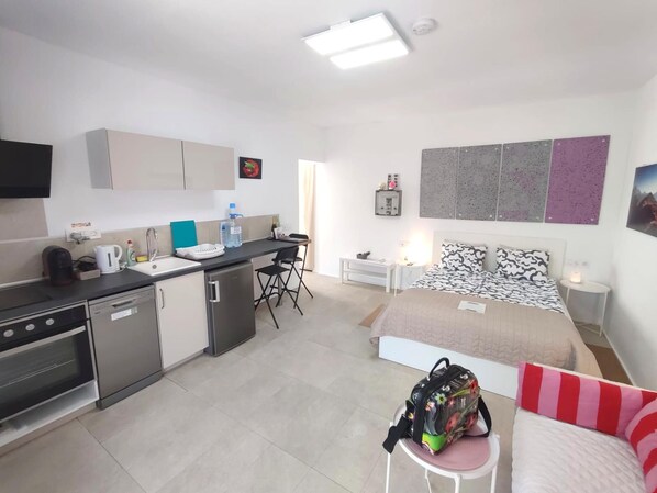 Une chambre d’hôtel compacte comprenant un coin cuisine, un lit avec une literie à motifs, un petit coin salon et un téléviseur fixé au mur.