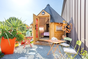Une petite cabane en bois, dont la porte est ouverte, laissant apercevoir un lit et une table sur laquelle se trouvent divers objets.