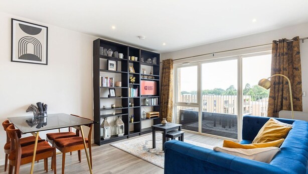 A modern living room with a glass table, dark wooden chairs, a blue sofa, and a large bookshelf.