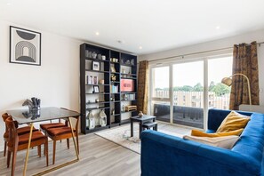 Een moderne woonkamer met een glazen tafel, donker houten stoelen, een blauwe bank en een grote boekenkast.