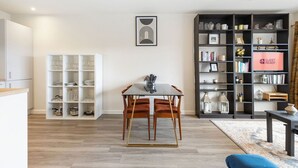 A modern living room with a dining area, featuring a glass table, wooden chairs, a bookshelf, and a 'GUEST READY' sign.