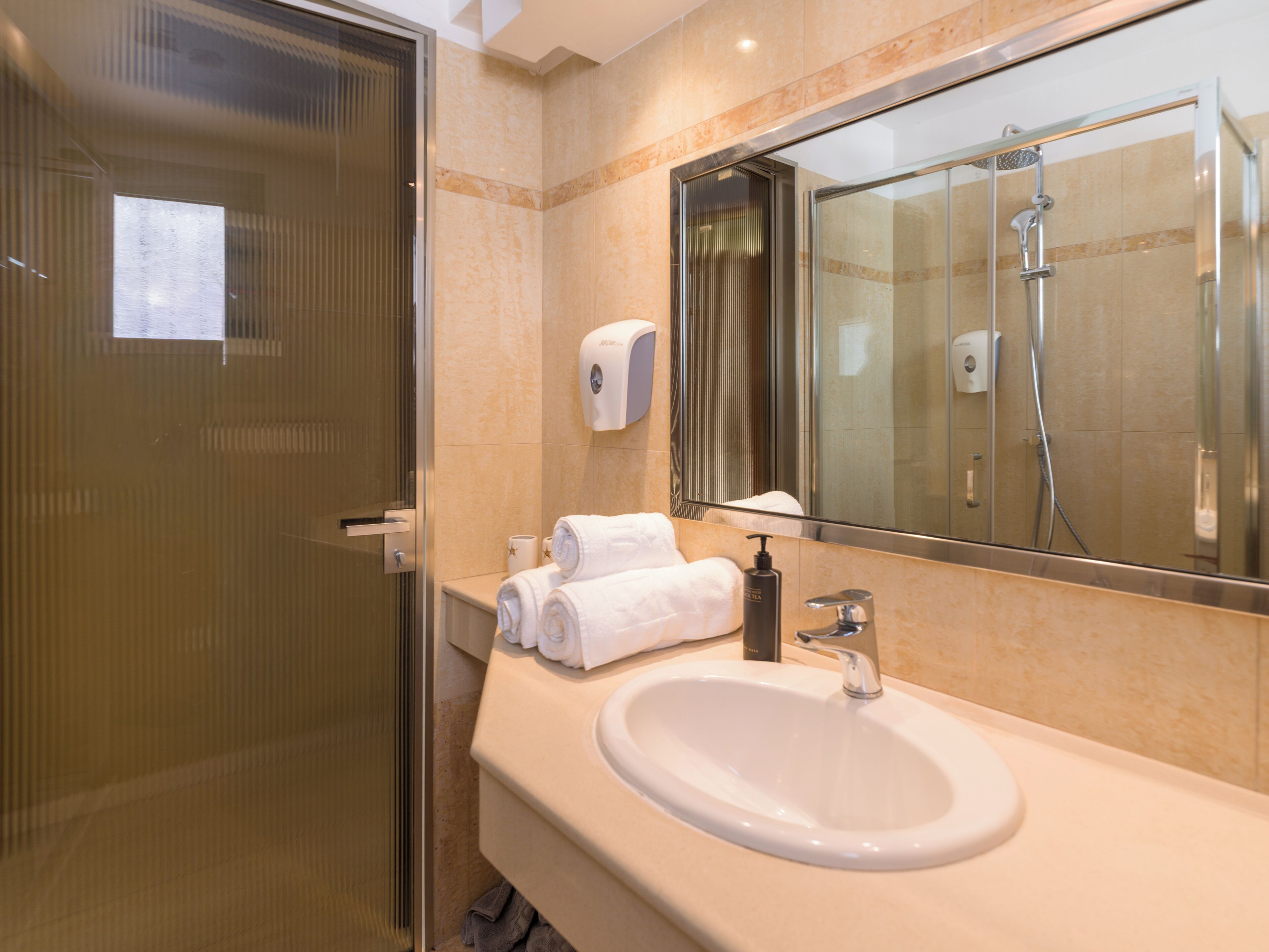 A bathroom with a glass shower enclosure, a sink with a mirror, and a towel dispenser.
