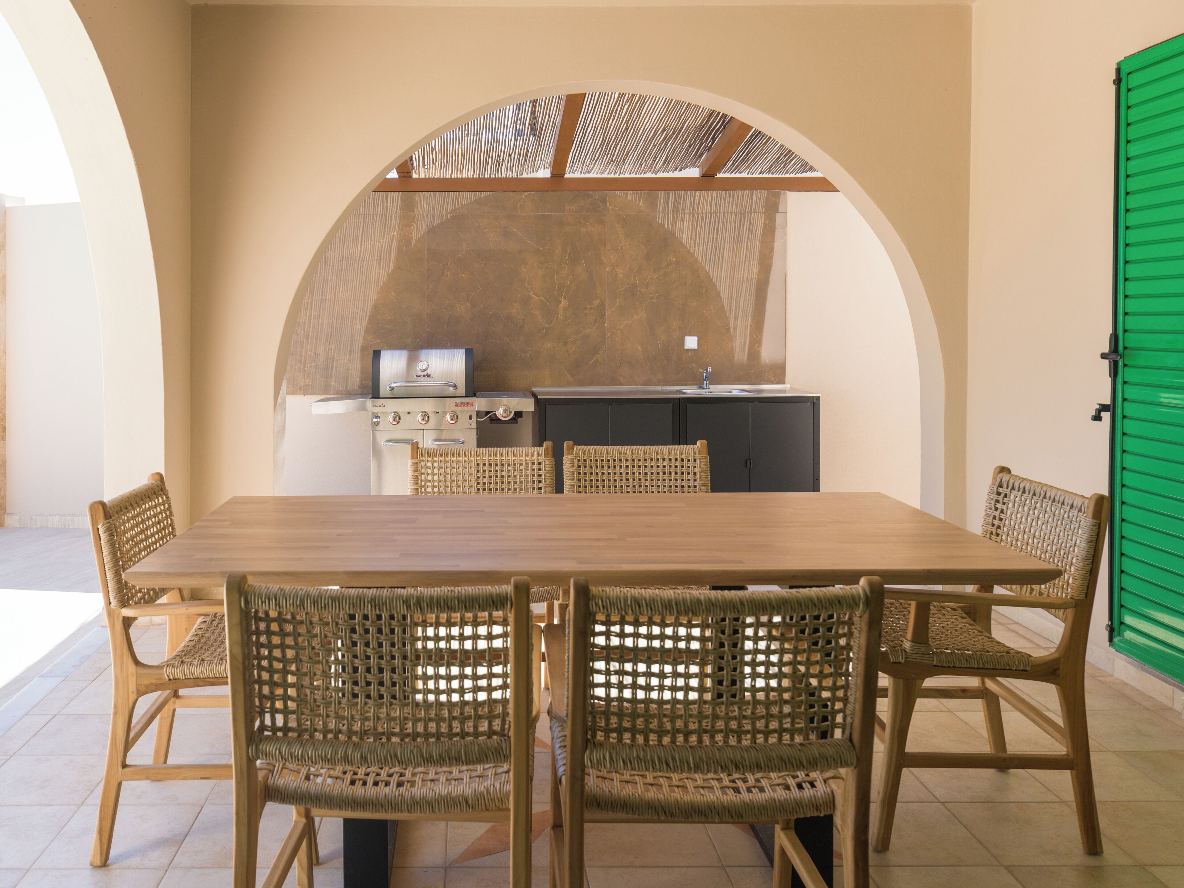 A dining area with a wooden table and wicker chairs, a kitchen with a grill, and a green door.