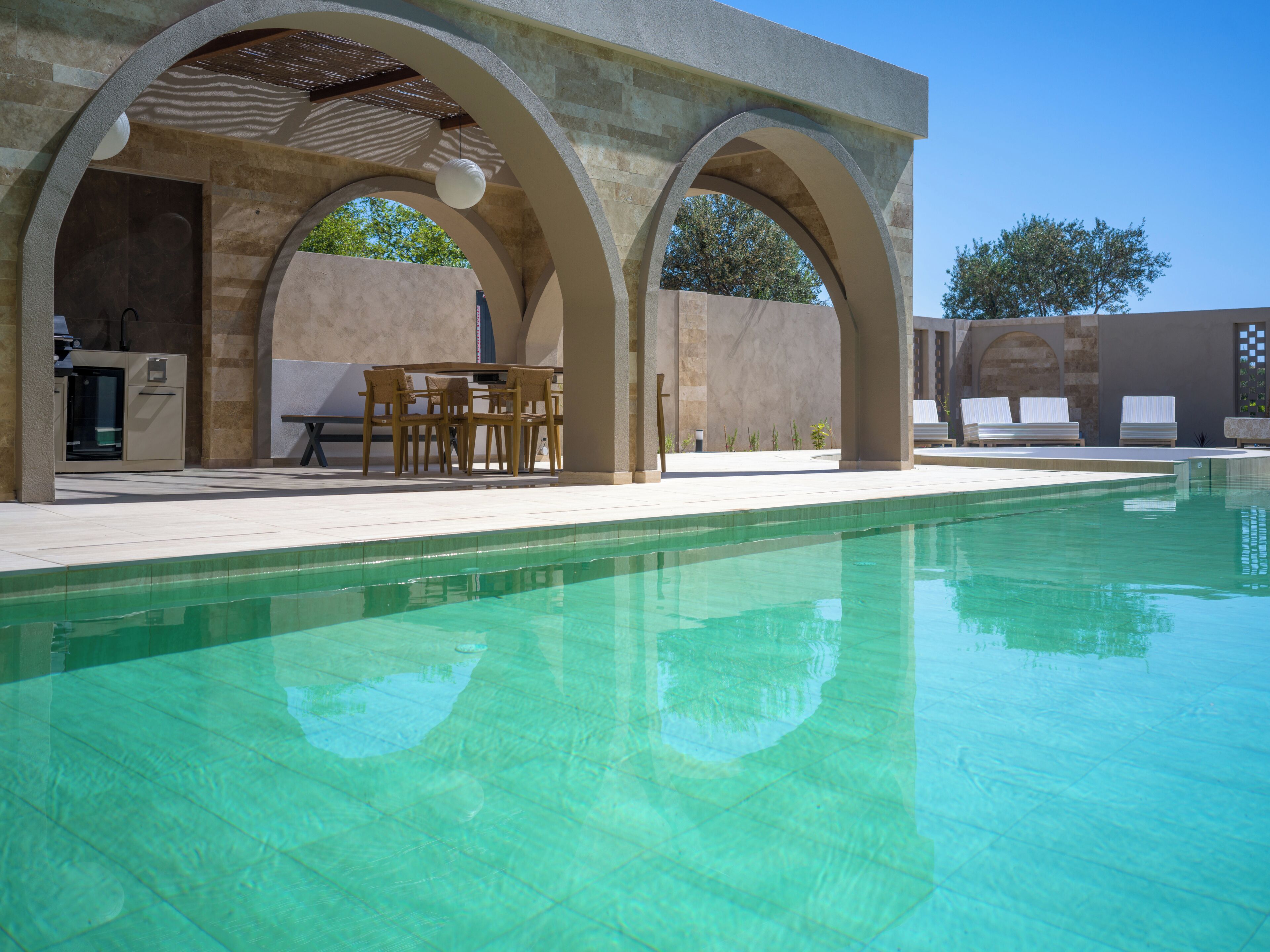 A swimming pool with clear water, surrounded by a covered patio area with seating and a table.