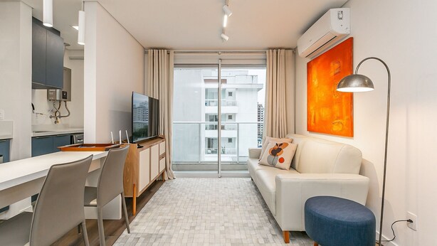 Apartamento moderno com cozinha, sala de jantar e sala de estar. Possui uma janela grande, televisão de tela plana e um sofá com um banquinho azul.
