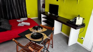 Una habitación compacta con un sofá rojo, una pequeña mesa de comedor con sillas, un televisor sobre un escritorio y un microondas en un estante.