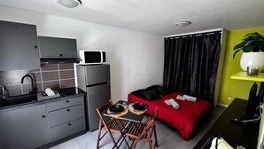 Una habitación compacta con kitchenette, una cama con ropa de cama roja, una pequeña mesa de comedor y una silla.