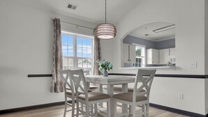 A dining area with a white table and chairs, a hanging light fixture, and a view of the kitchen through the window.