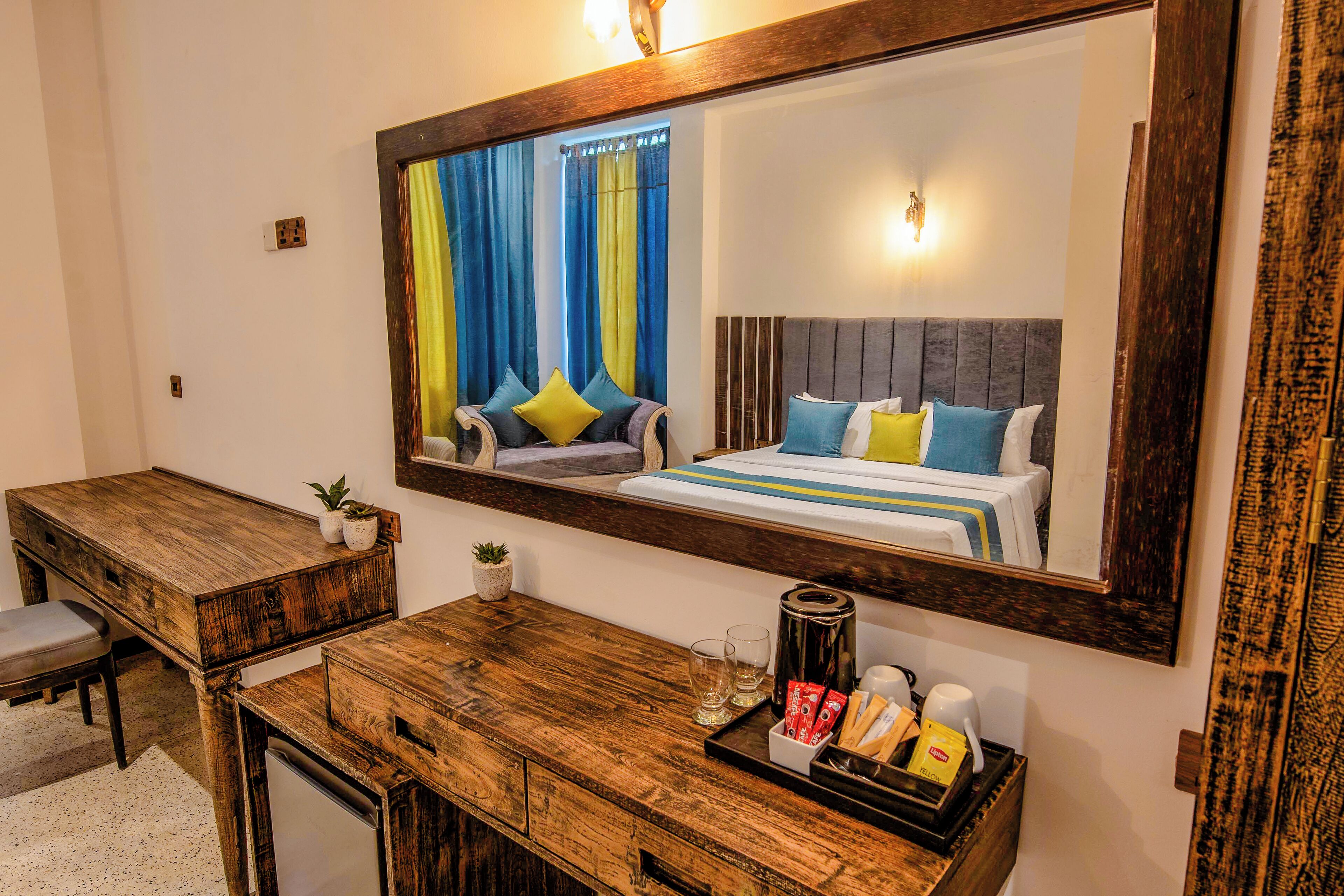 A hotel room with a wooden desk, a bed with blue and yellow bedding, a large mirror, and a small potted plant.