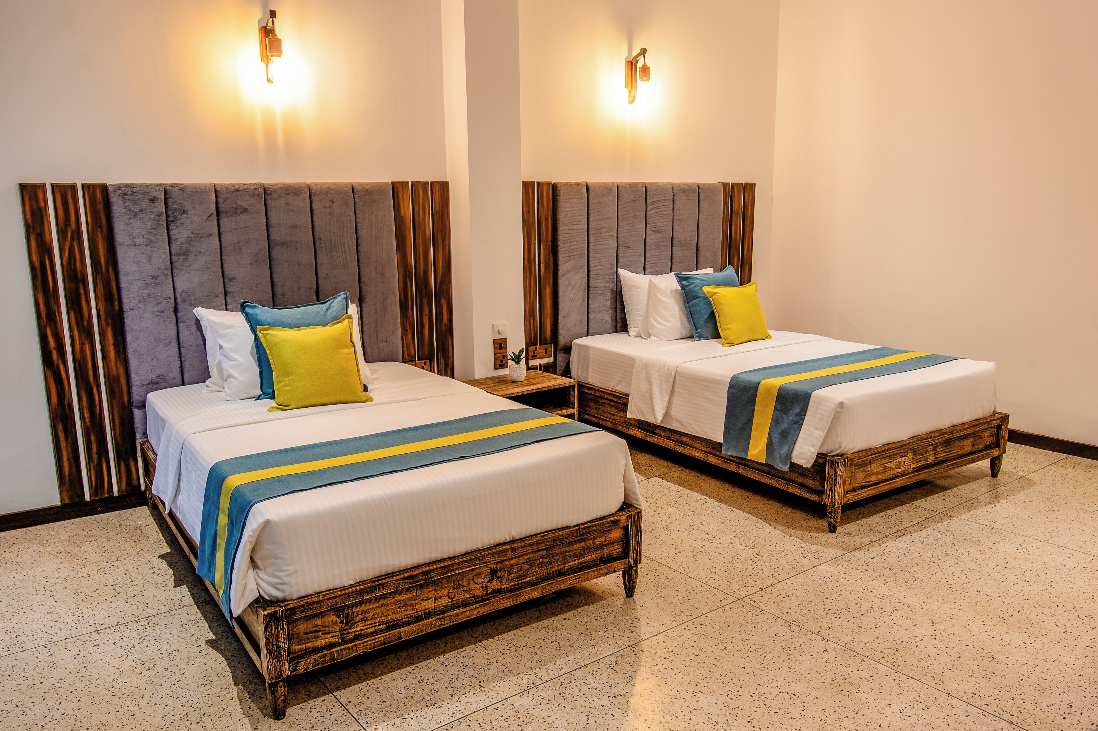 Two wooden beds with white linens and blue and yellow striped bedspreads, flanked by wooden headboards and wall-mounted lights.
