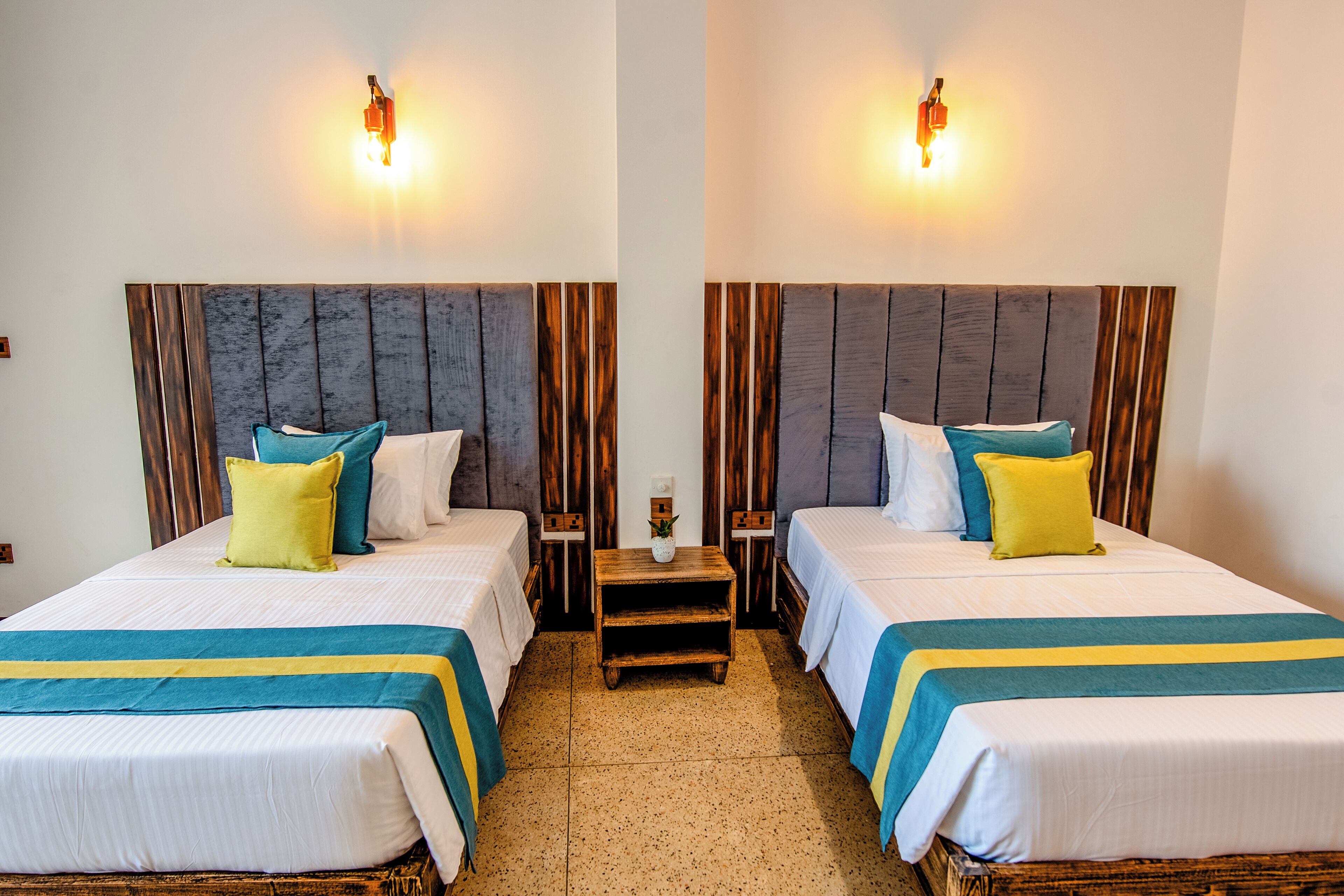 Ein Hotelzimmer mit zwei Betten, beide mit Holz Kopfteilen, weißer Bettwäsche und Türkis- und Gelbtönen. Zwei Nachttische mit Lampen und eine kleine Holzablage zwischen den Betten.