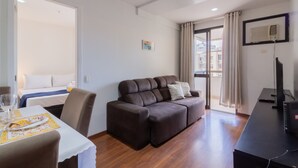 A modern living room with a brown sofa, a dining table with chairs, a TV, and a balcony with a view of buildings.