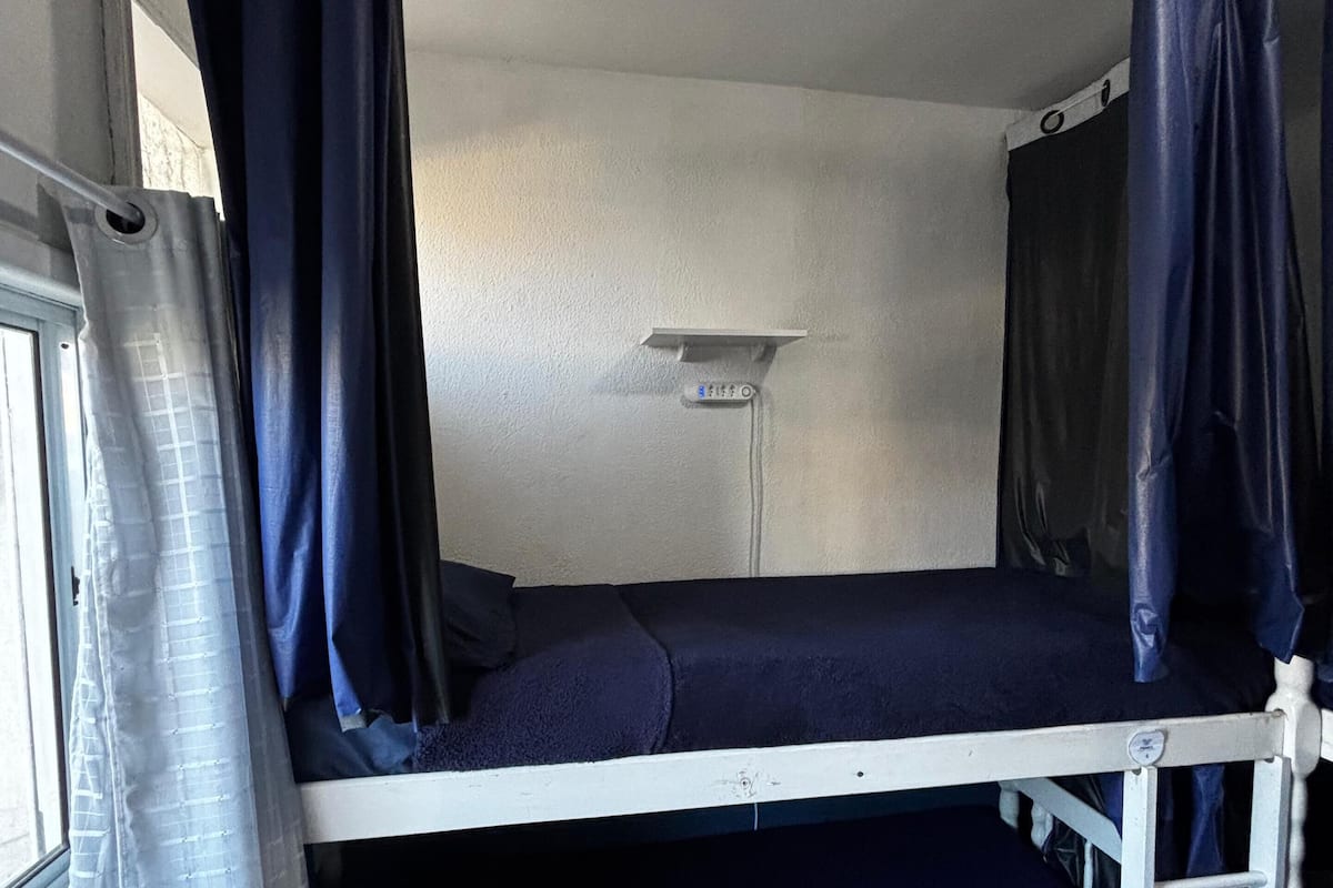 Una cama litera con ventana y cortinas, un estante pequeño y una lámpara fijada a la pared.