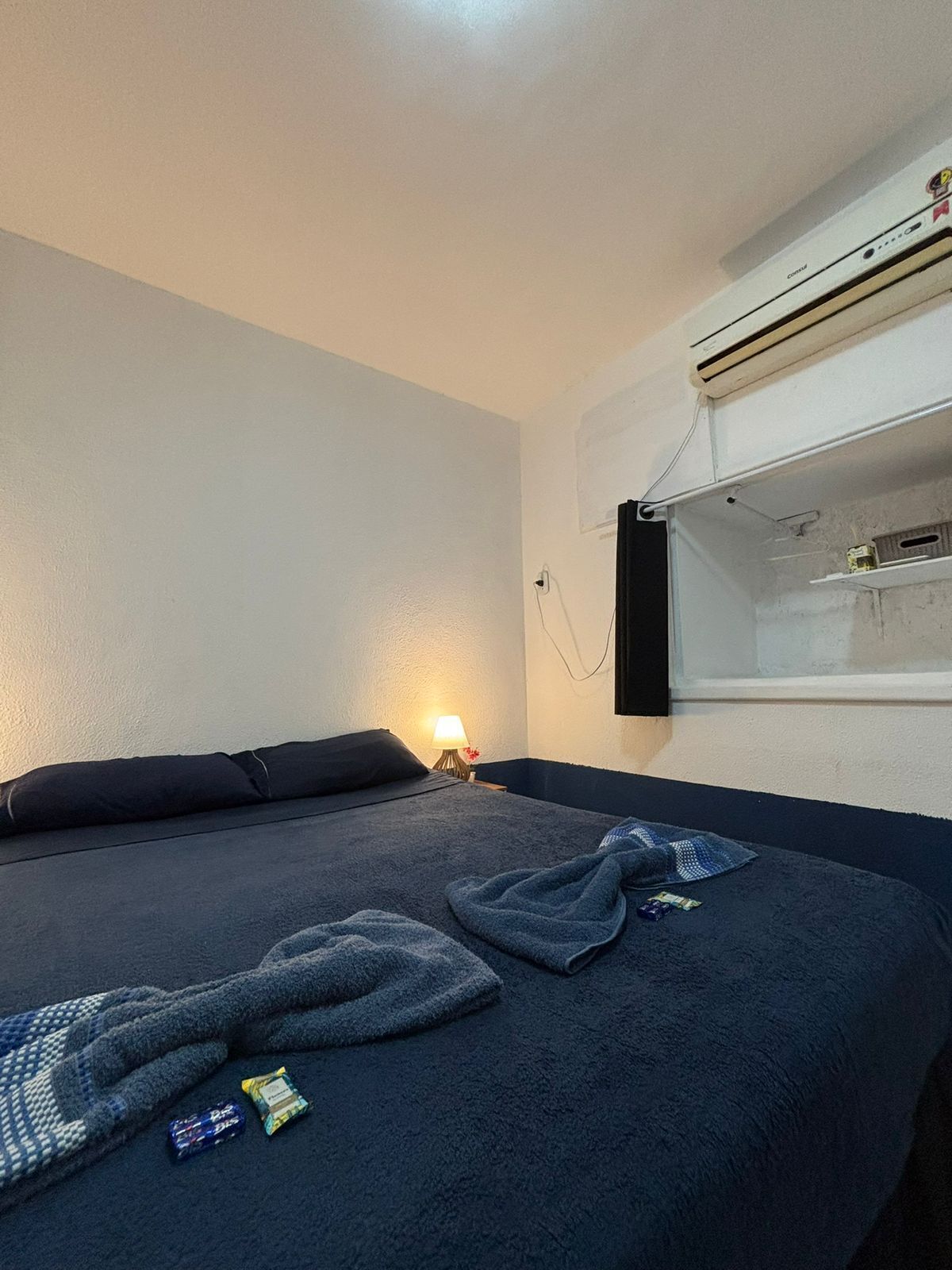 A single bed with a dark blue blanket and pillows, a bedside lamp, and a wall-mounted air conditioning unit.
