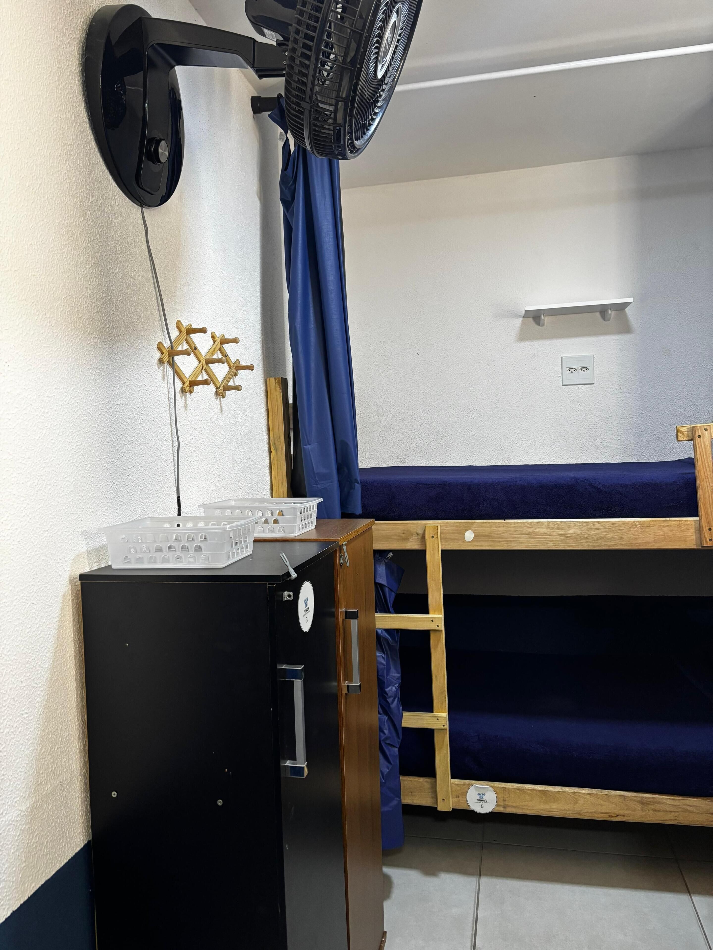 Quarto pequeno com beliche, secretária compacta com computador, ventoinha fixada na parede e tomada.