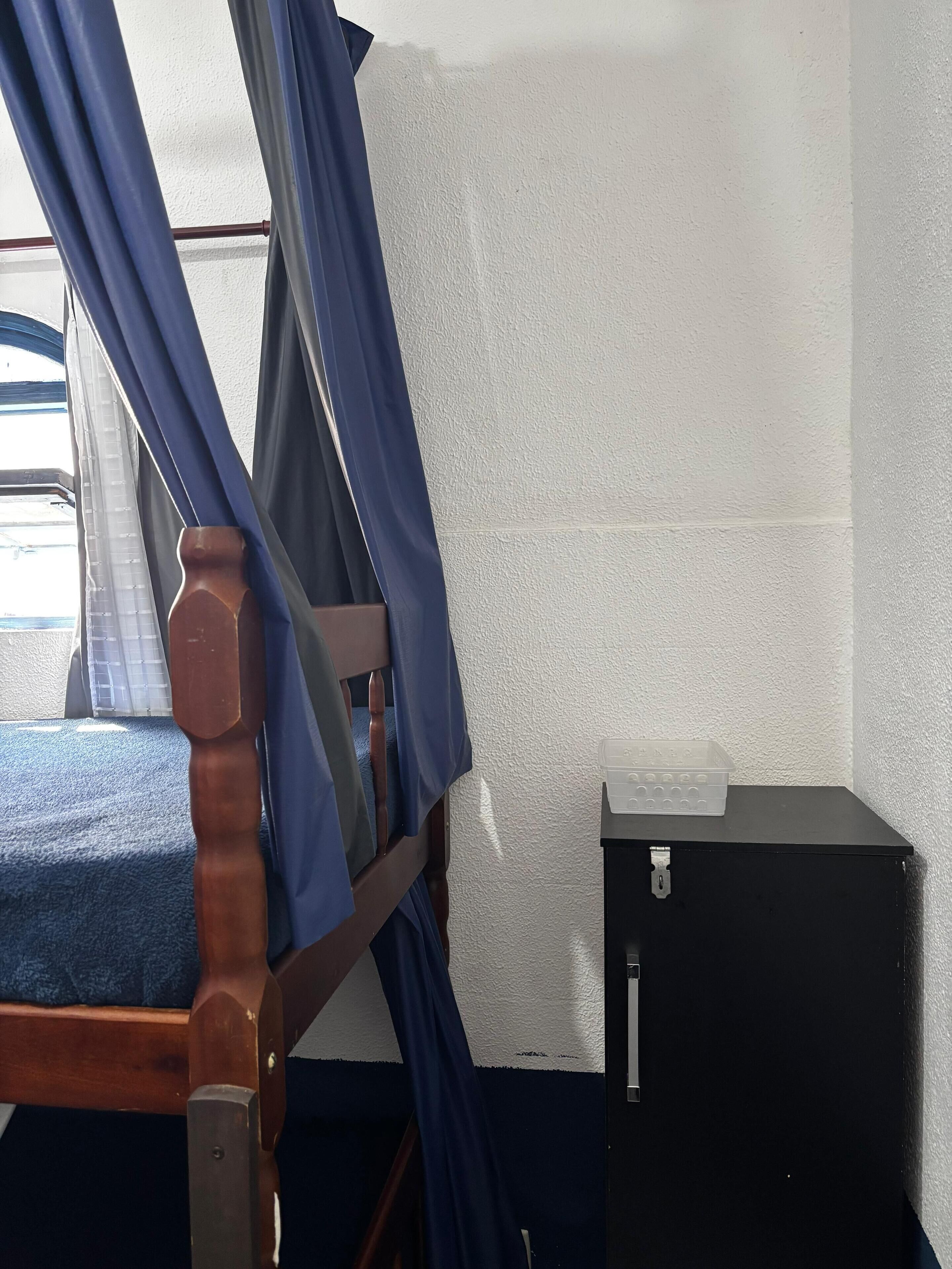 Una habitación con una cama, una ventana con persianas, un calentador de pared y un mueble negro pequeño.