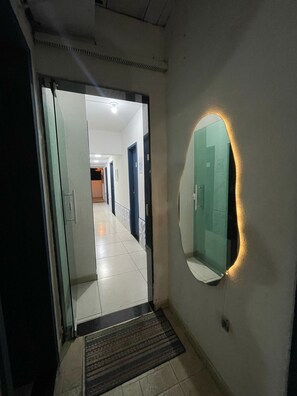 A hallway with a glass door leading to a room, a round mirror with a light, and a tiled floor.