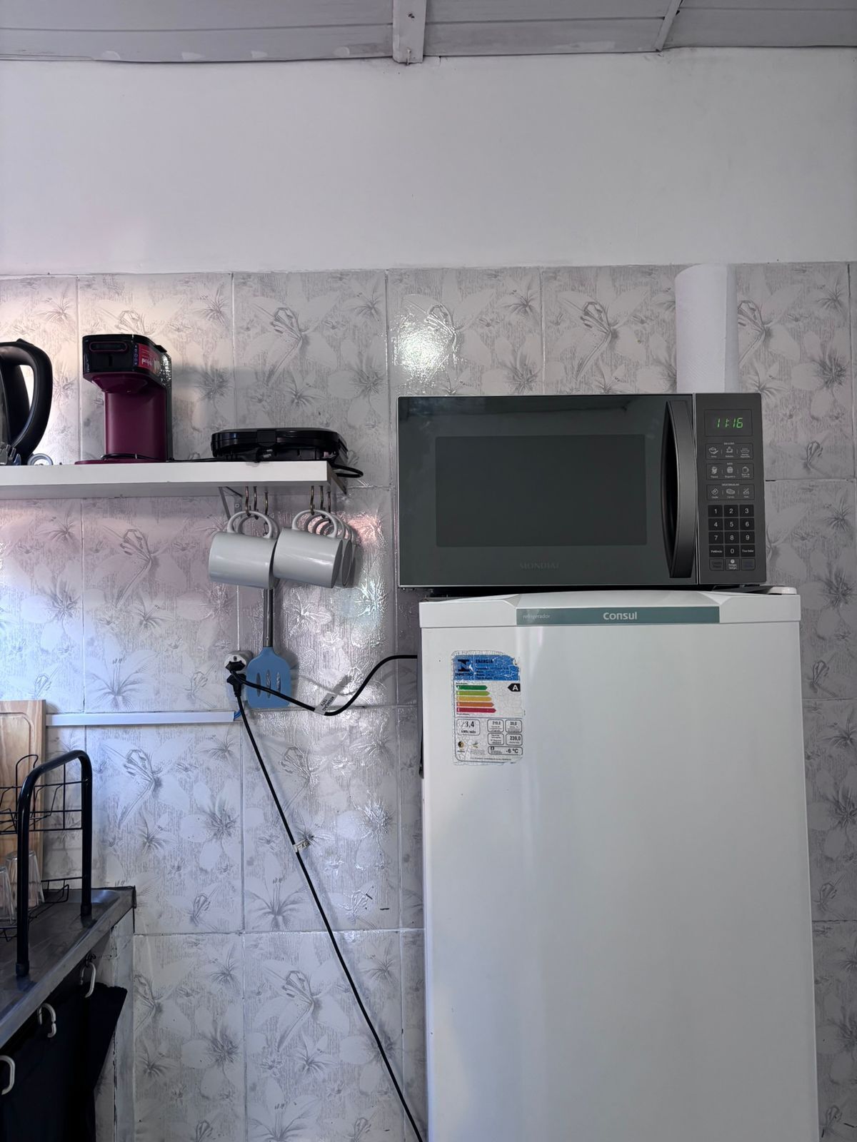 A kitchen with a microwave, a refrigerator, and a kettle on a shelf.