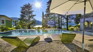 A swimming pool with lounge chairs and an umbrella, surrounded by trees and a mountain in the background. - Elegant Historic Villa with Pool, Jacuzzi & Lake Views in Lierna (Lierna)