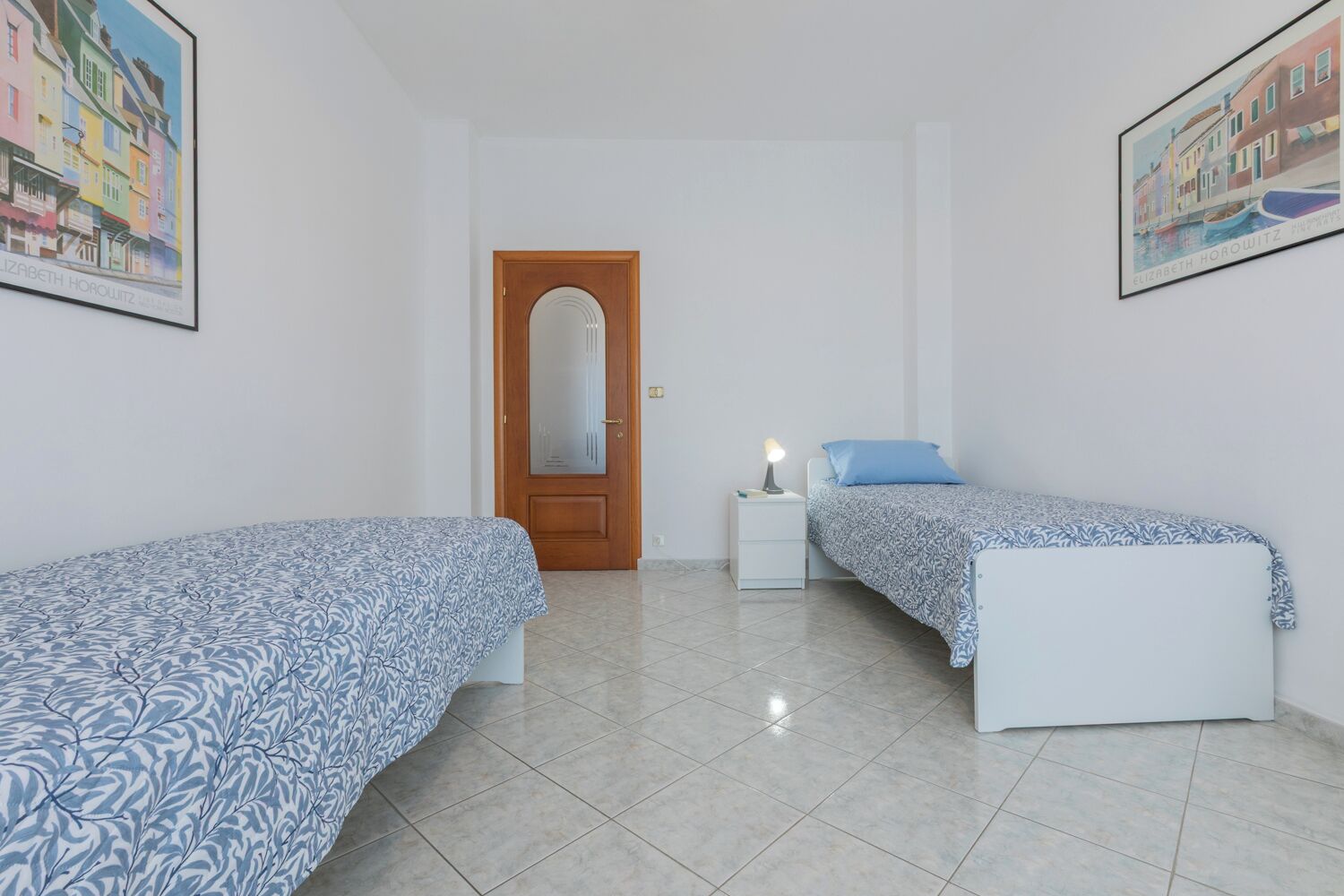 Un dormitorio con dos camas, una puerta de madera, una mesita de noche con una lámpara y dos cuadros enmarcados en las paredes.