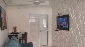 Uma sala de estar com um sofá azul, uma TV fixada na parede, um ventilador de teto e uma parede com padrão.