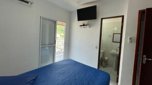 Quarto com cama, ventilador de teto, televisão montada na parede e banheiro visível através de uma porta aberta.