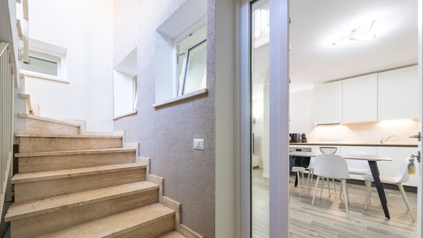 Un intérieur moderne avec un escalier menant à une salle à manger, une cuisine dotée de meubles blancs et une porte-fenêtre coulissante.
