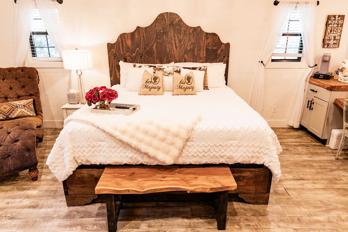 Una camera da letto con una testiera in legno, una coperta bianca, due cuscini con la scritta 'Her Majesty' e 'His Majesty', un comodino con una lampada, una panca in legno e una finestra con tende.