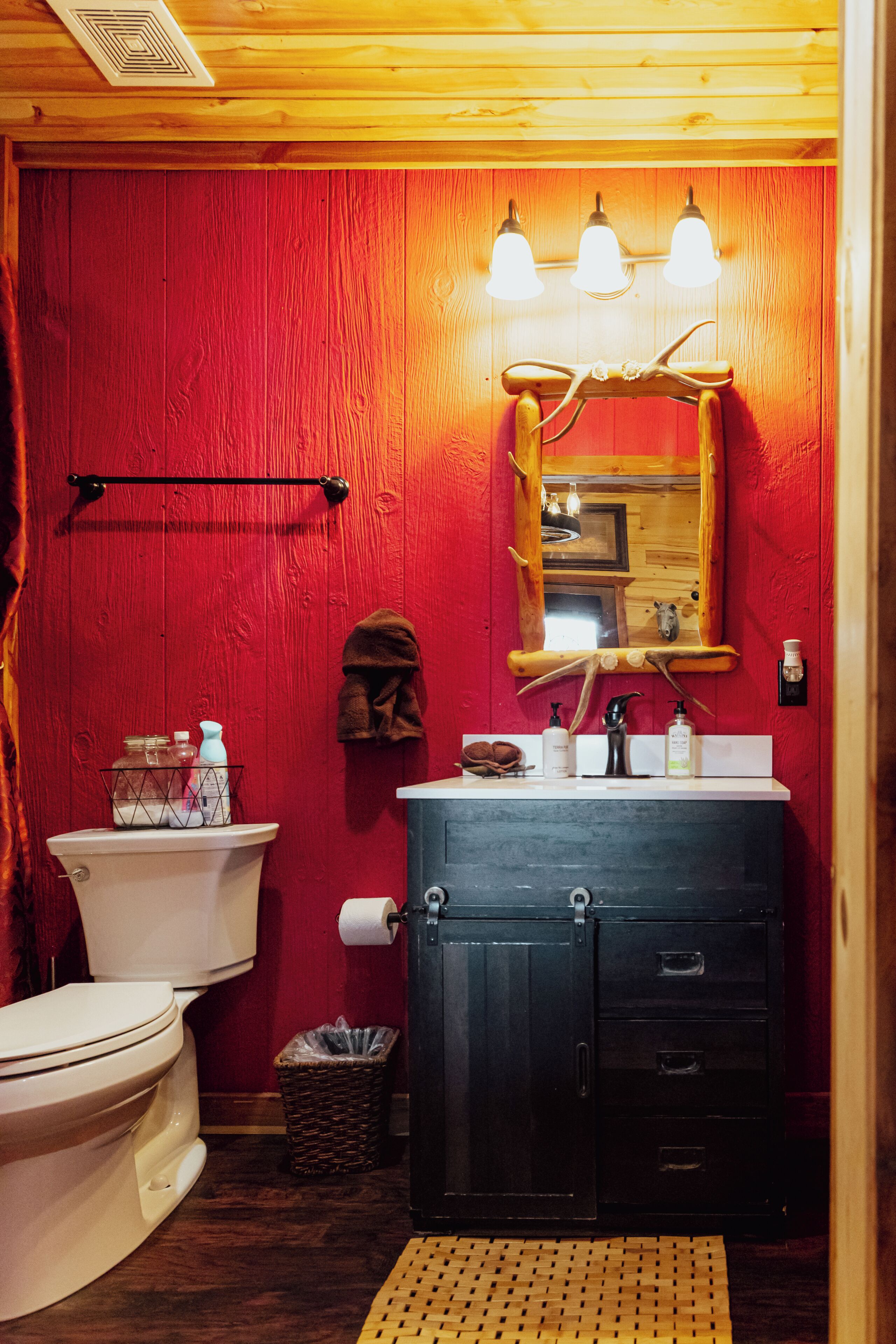 Une salle de bain avec un mur rouge, un plafond en bois, un meuble noir, des toilettes blanches et un miroir orné d’une tête de cerf.