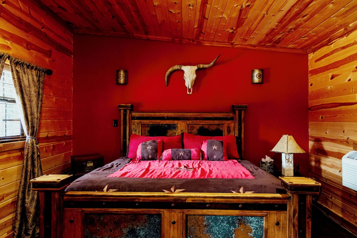 Camera di una baita in legno con un letto, pareti rosse, soffitto in legno e un teschio di mucca appeso alla parete.