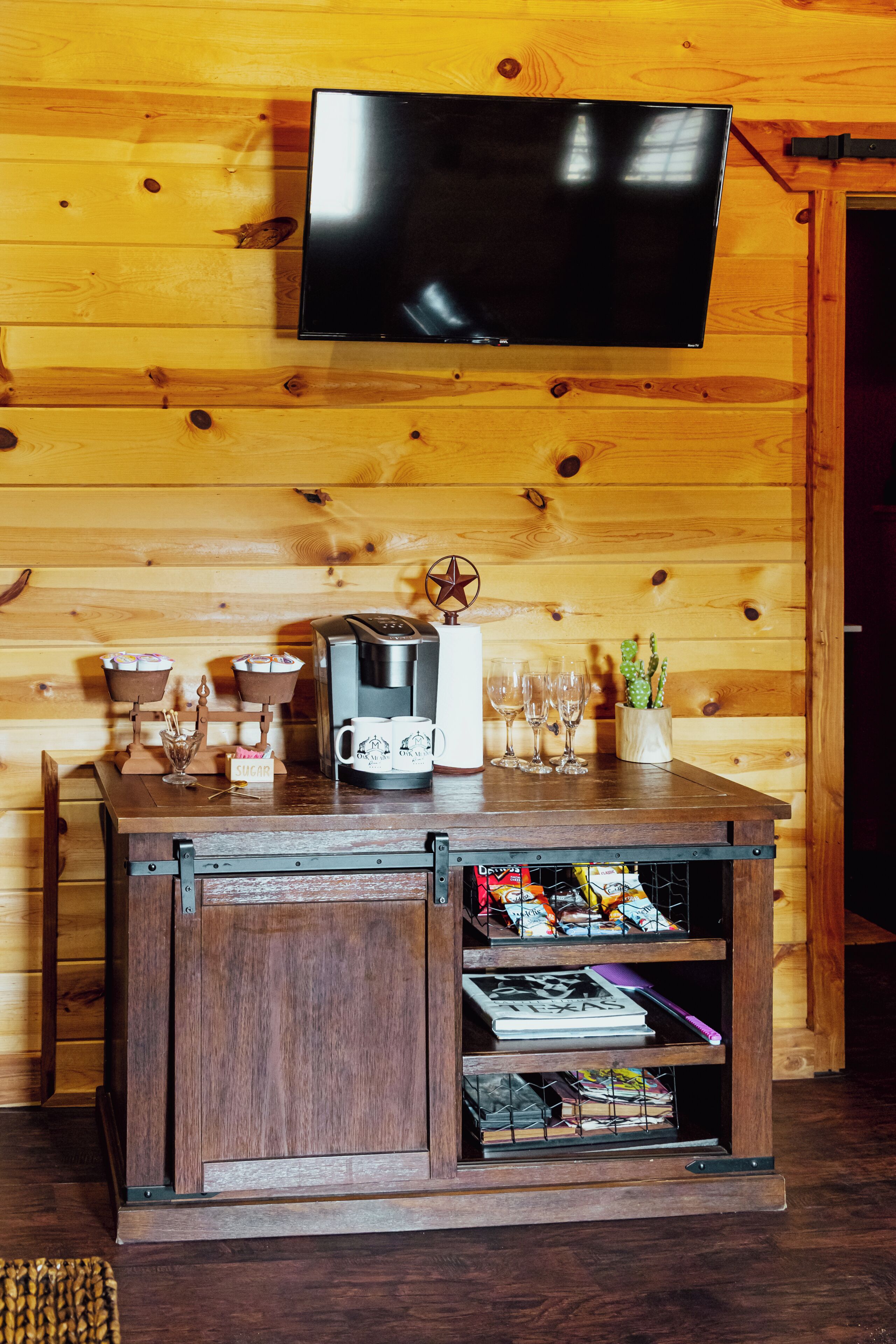 A wooden interior with a TV mounted on the wall, a coffee machine, cups, glasses, and a cactus in a planter.