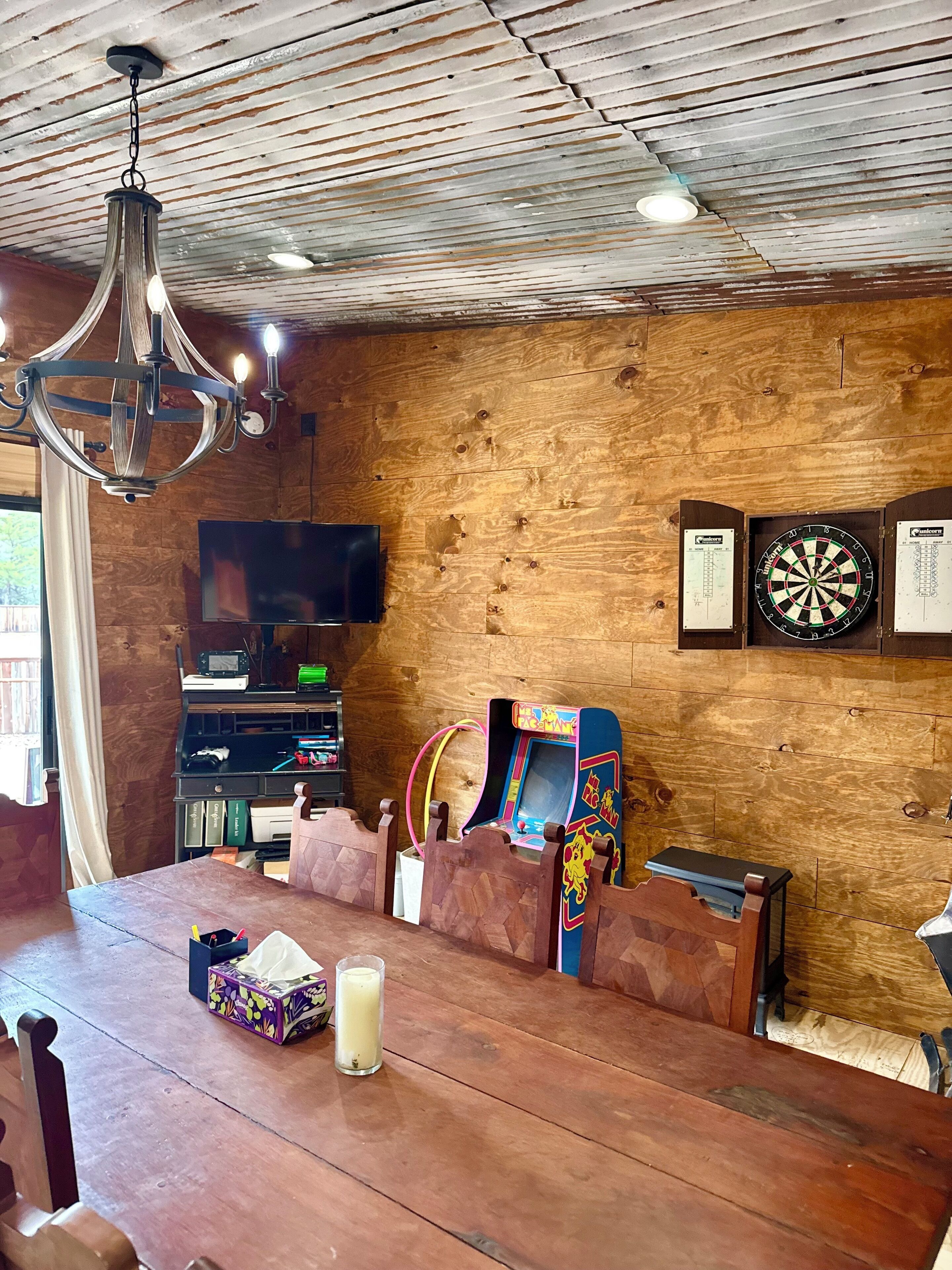 A cozy room with a wooden ceiling, a chandelier, a dartboard, a dartboard stand, a television, and a gaming console.