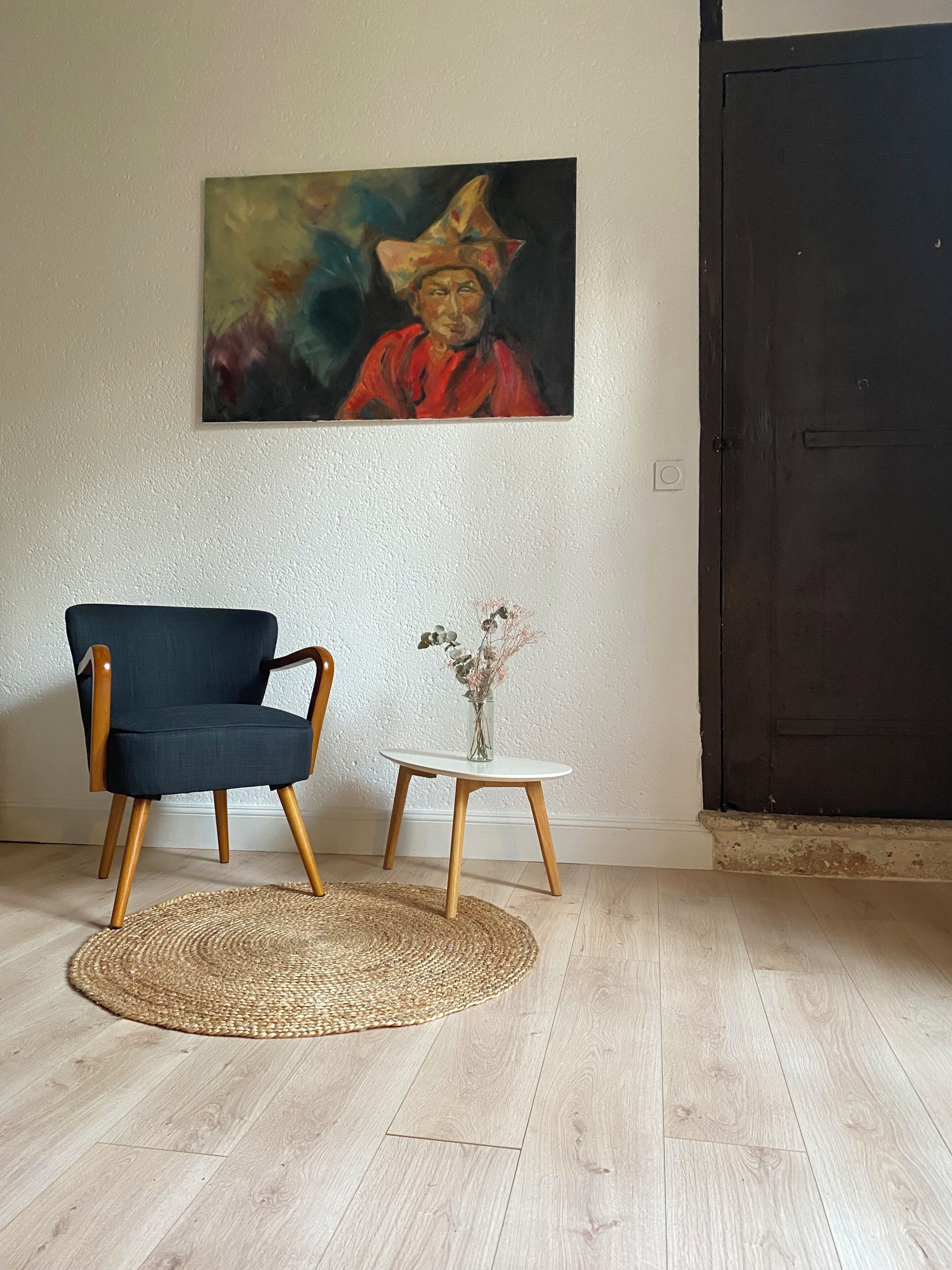 Une pièce avec un fauteuil bleu foncé, une petite table blanche sur laquelle se trouve un vase, un tapis tissé et un tableau accroché au mur.