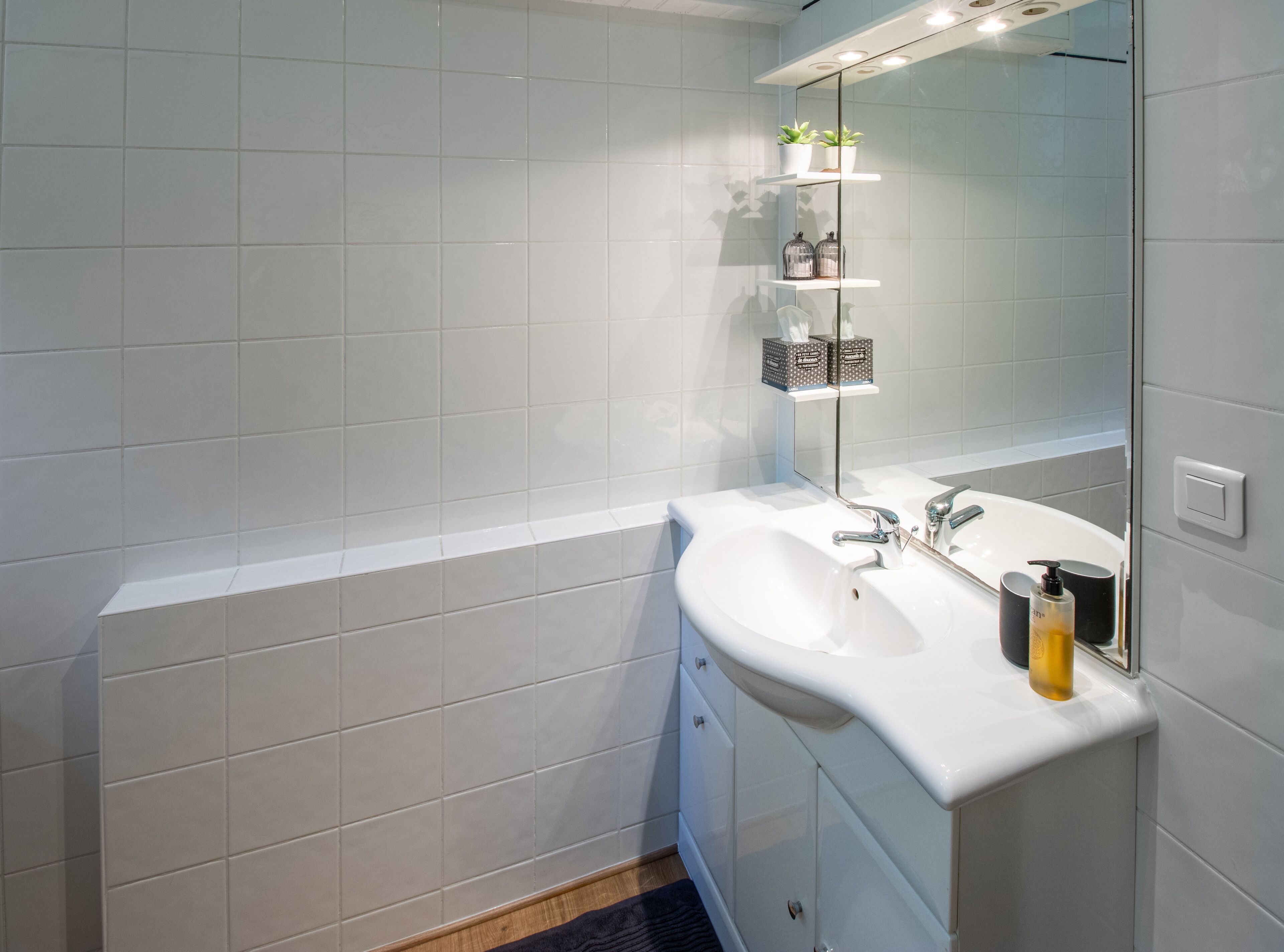 Ein Badezimmer mit weißen Fliesen, einem weißen Waschbecken, einem Spiegel und einem Ablagefach für Pflegeprodukte.
