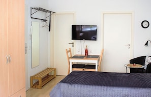 A bedroom with a bed, a TV mounted on the wall, a wooden table with two chairs, a small round table with a lamp, and a wardrobe.
