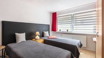 Ein Hotelzimmer mit zwei Betten, einem Nachttisch mit Lampe, einem Fenster mit Jalousien und Blick ins Freie.