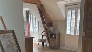 Habitación con suelo de madera, un cuadro sobre caballete, una silla, un armario de madera y una ventana con vistas a edificios de piedra.