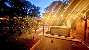 Un jacuzzi bajo una pérgola con luces colgantes, rodeado de árboles y una zona de patio.