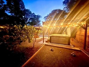 Een hottub onder een pergola met lichtjes, omringd door bomen en een terras.