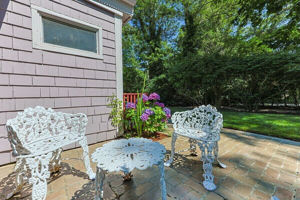 Eine Terrasse mit zwei verzierten weißen Metallstühlen und einem Tisch, ein rosafarbenes Haus und ein Garten mit violetten Blumen.