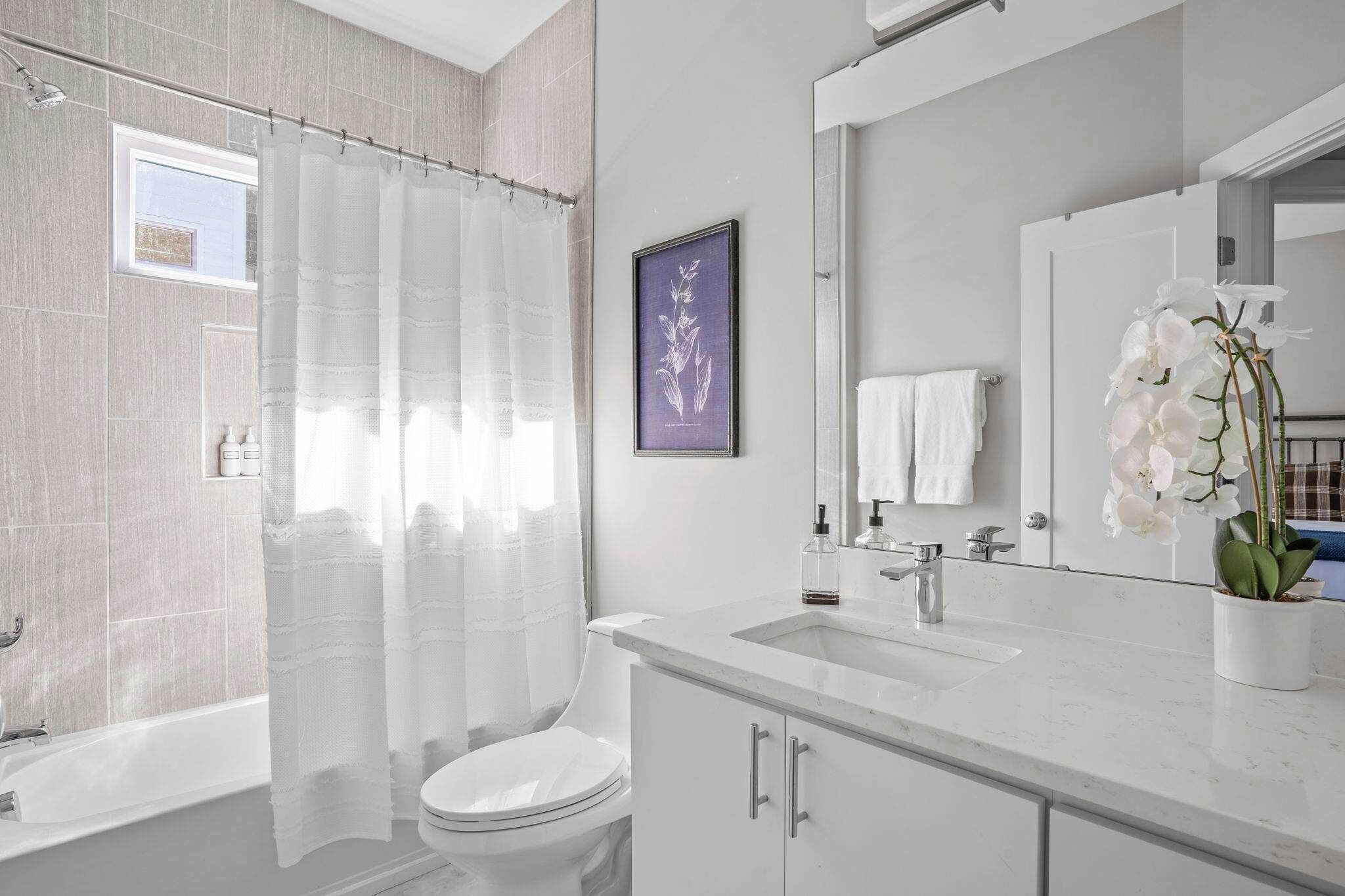 Un baño moderno con bañera, inodoro y lavamanos. Hay una cortina de ducha, un cuadro en la pared y una orquídea en maceta sobre el mostrador.