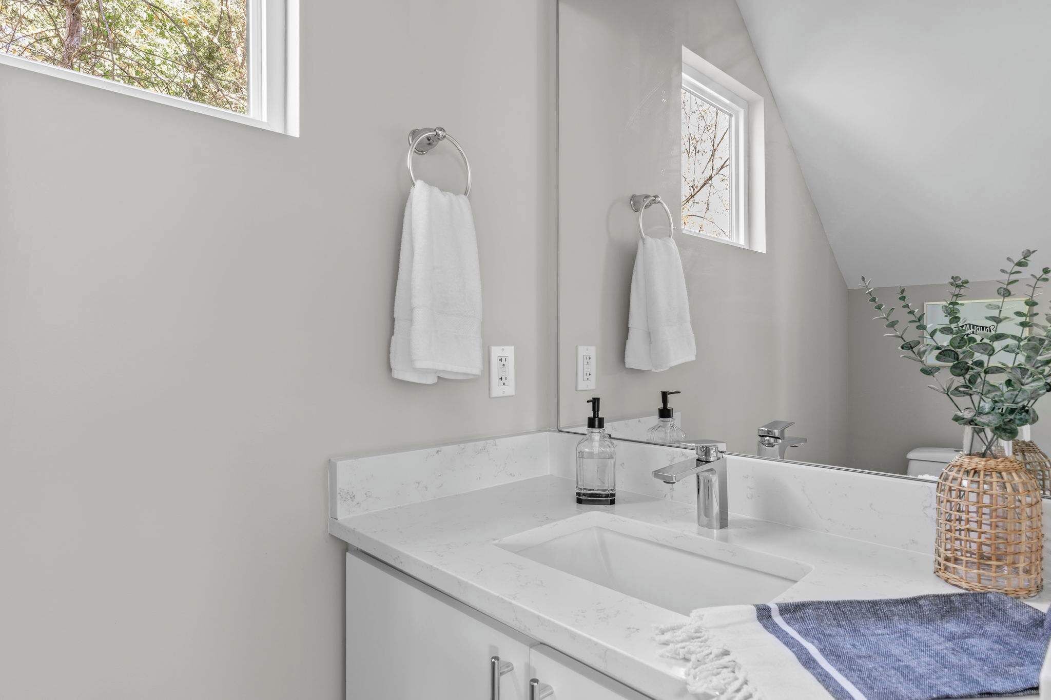 A modern bathroom with a large mirror, two towel rings, a sink, and a window.